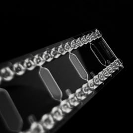 A fluidic chip by microfluidic ChipShop infront of a black background. The chip is translucent and has four rhombic chambers with one inlet and one outlet each. They are accessible via mini luer connections.